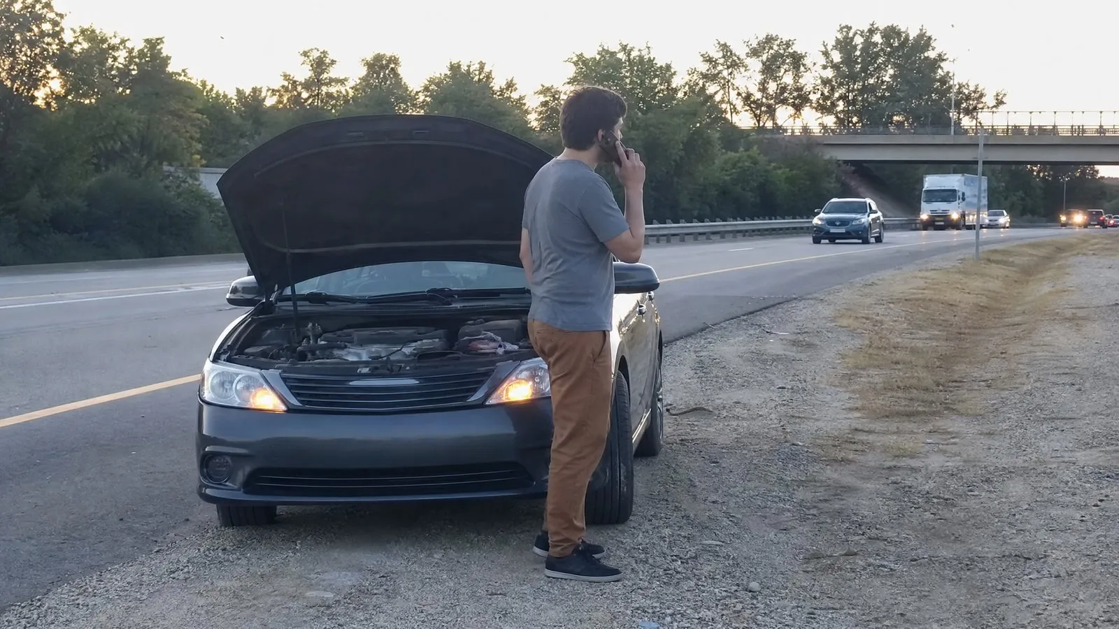 A driver stands at the open hood of an ordinary sedan parked on a highway shoulder with hazard lights on, holding a phone to call for help in soft evening light.