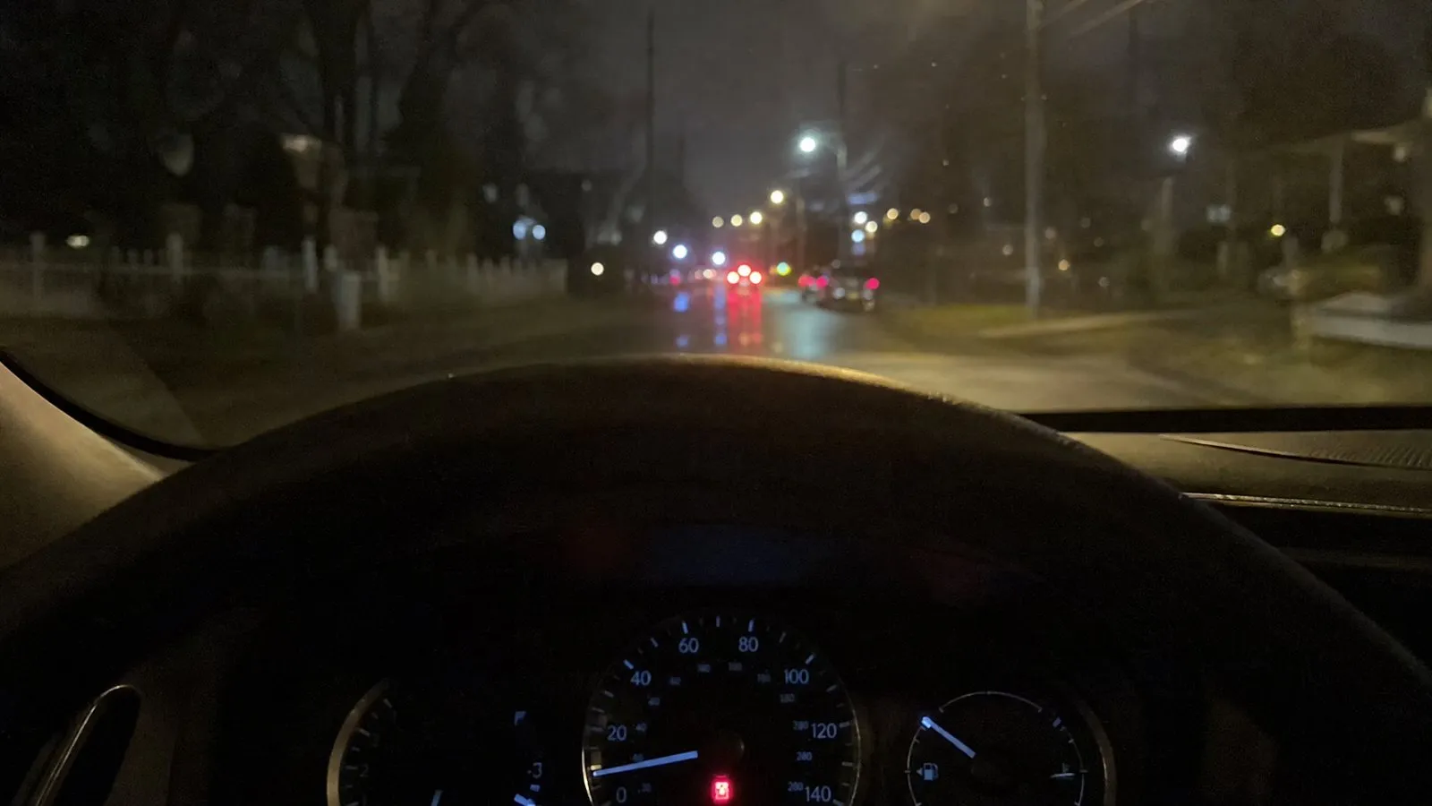 Nighttime view from a driver's seat looking at the dashboard where a red indicator glows, with blurred street lights through the windshield, shot on a phone.