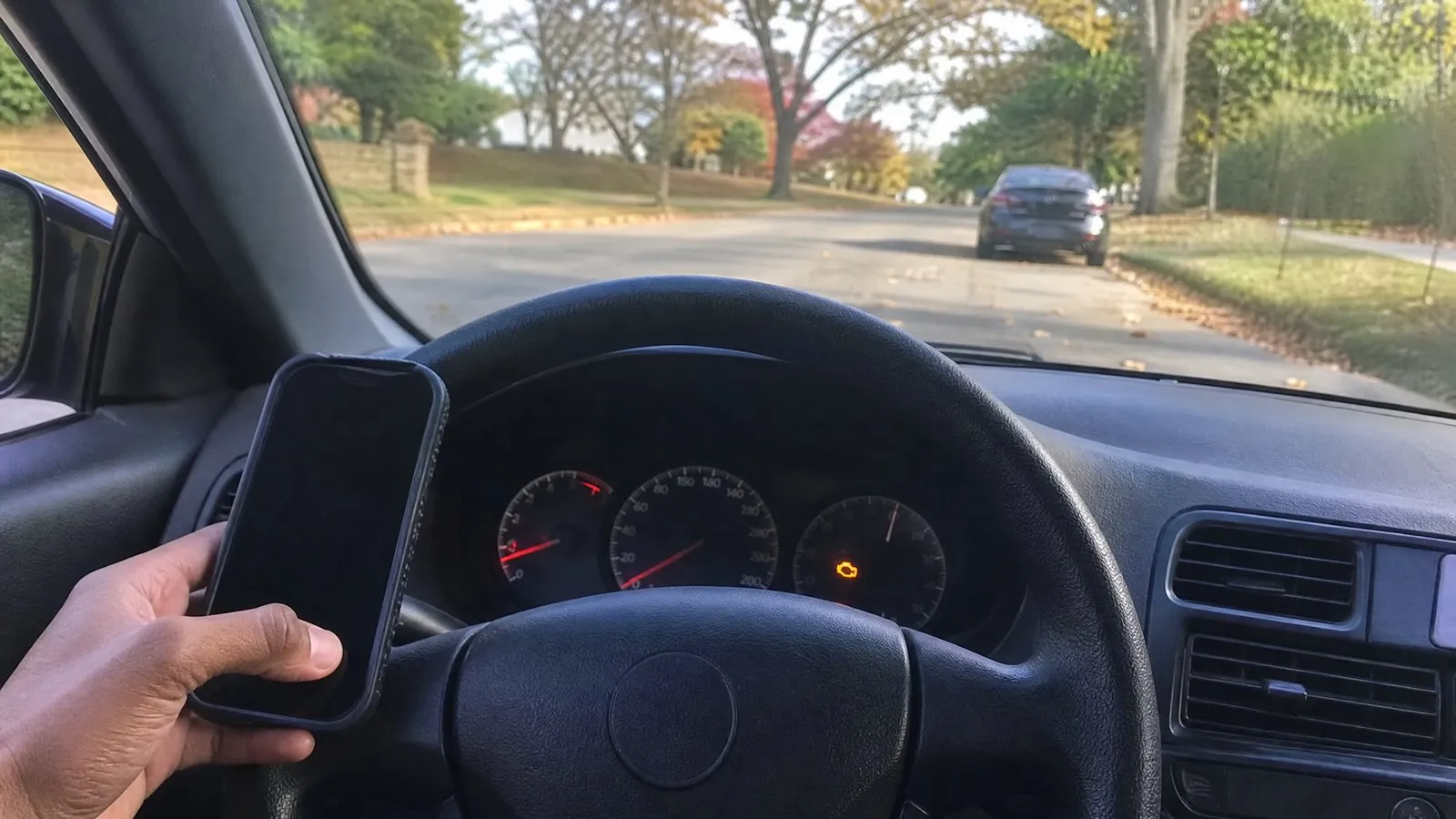 Driver's-seat view of a dashboard with an amber warning light glowing while a driver holds a phone to look up information, car parked at the roadside in natural daylight.
