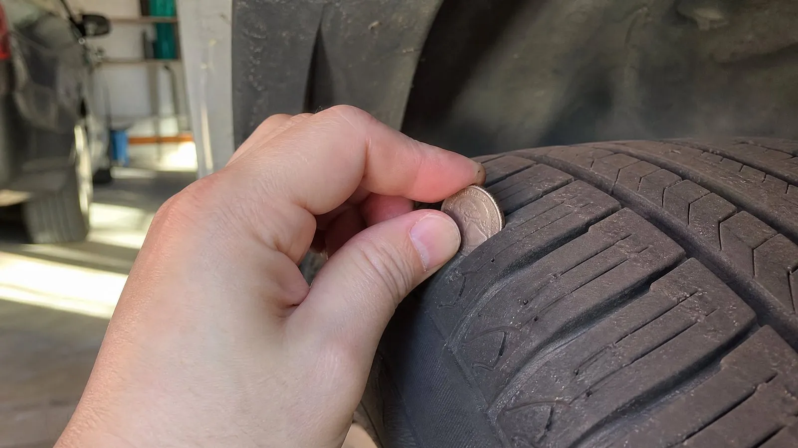 Close-up of a tire tread with a hand inserting a coin upside down into a groove to check depth, showing normal wear under side lighting in a realistic photo.
