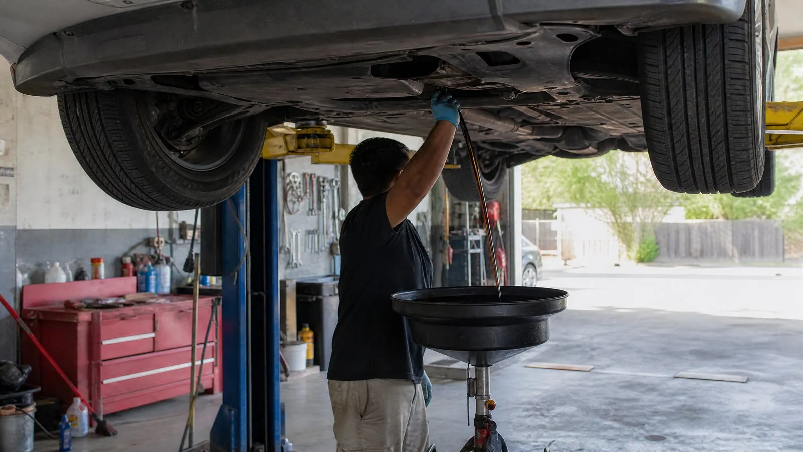 A family sedan raised on a lift in an independent repair shop while a technician drains old oil into a pan, with a tool wall and natural daylight in a real working garage.